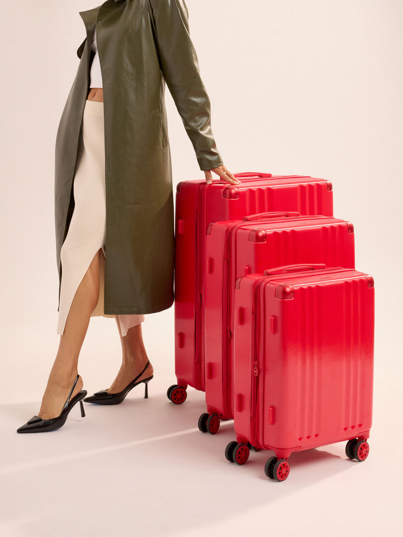 Model standing besides CALPAK Ambeur Large Luggage, Ambeur Medium Luggage and Ambeur Carry-On Luggage lined up tallest to shortest in metallic red, part of 3-piece luggage set