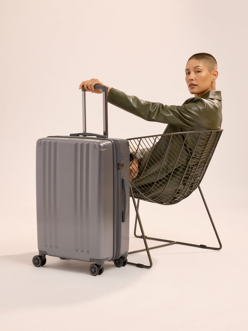 Model sitting besides CALPAK Ambeur Medium Luggage while holding telescopic trolley handle in metallic gunmetal gray