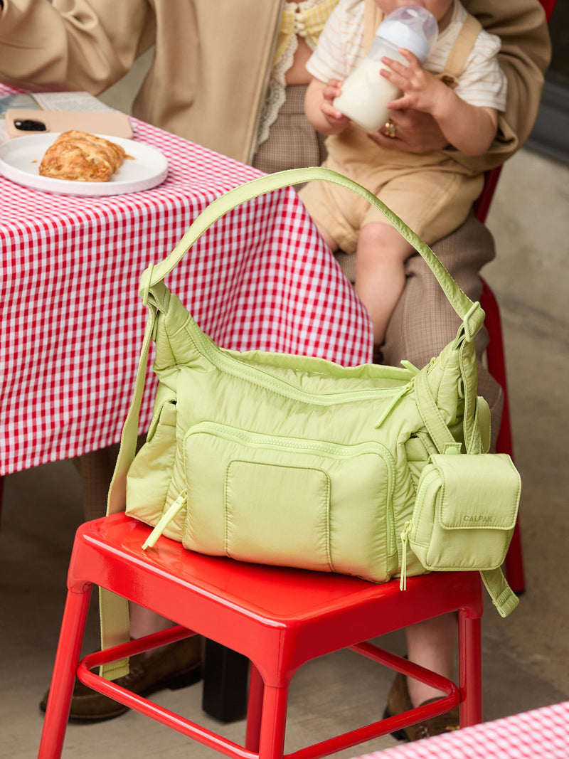 CALPAK Crossbody Diaper Bag in brown light green Lime on a red stool with a mom and baby in the background