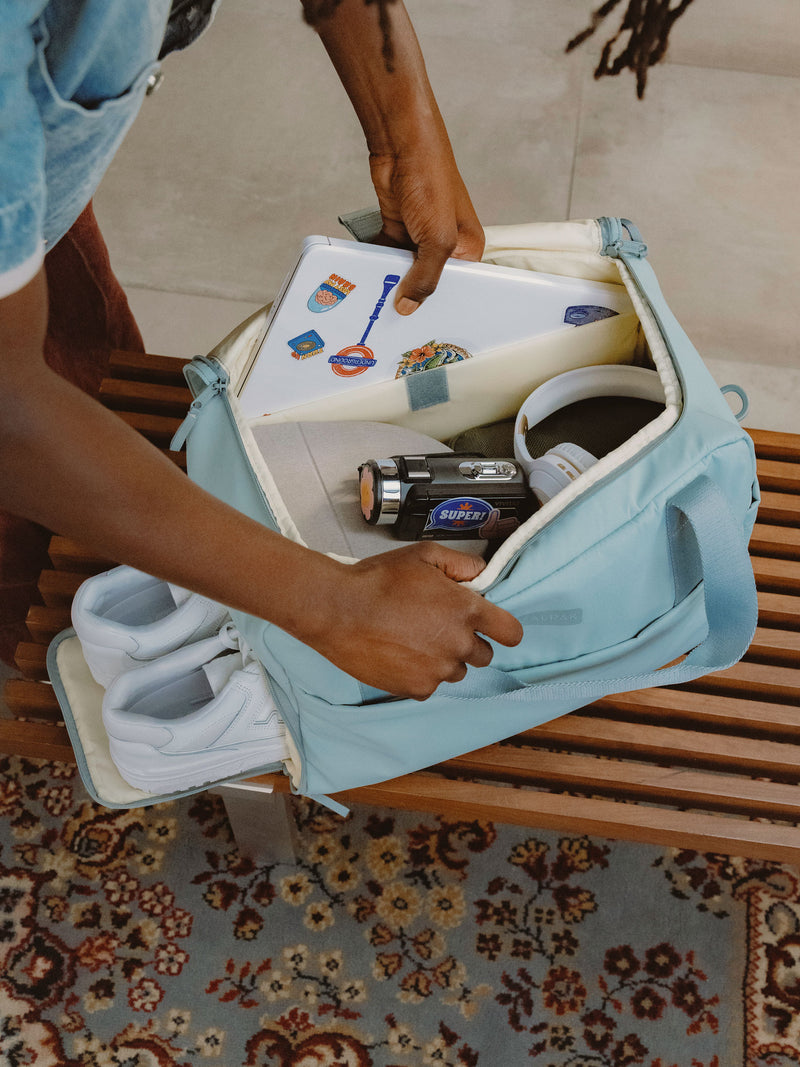 Model placing laptop within padded laptop sleeve of blue CALPAK Connect Laptop Duffel with other belongings inside