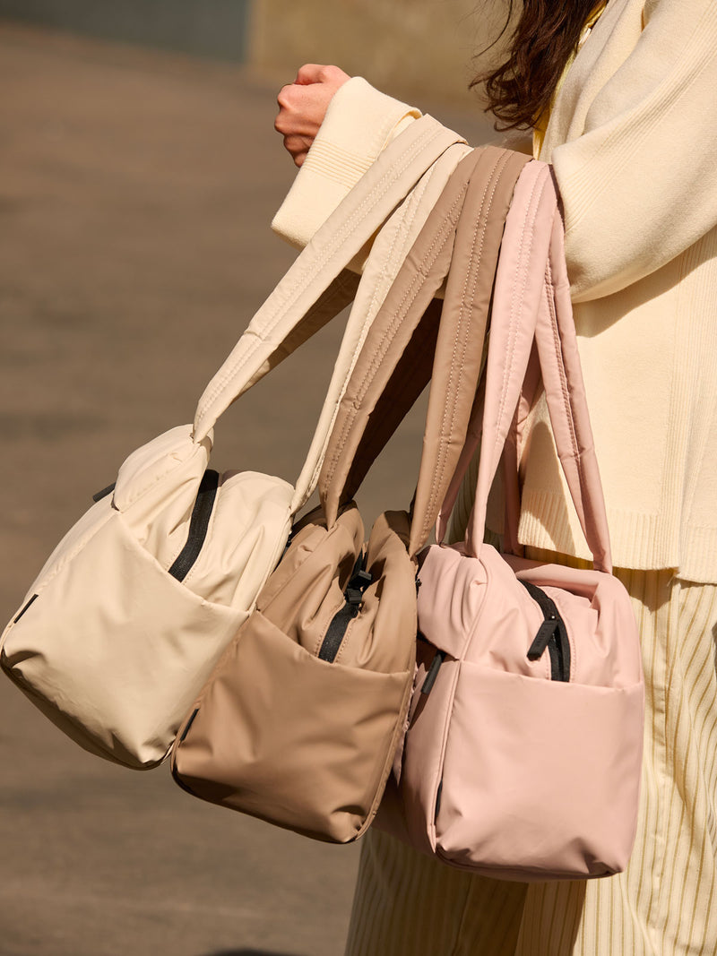 CALPAK Mini Duffel in brown Chocolate, white Oatmeal, and pink Rose Quartz with puffy exterior, interior and exterior pockets, and padded shoulder straps being held by a model's forearm