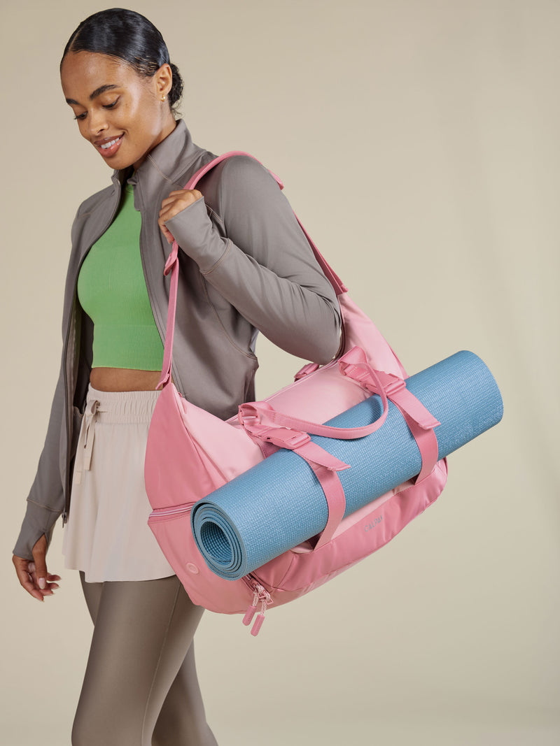 Model carrying Fit Gym Bag over shoulder with yoga mat secured to bag by the adjustable yoga mat straps on the exterior of the duffel