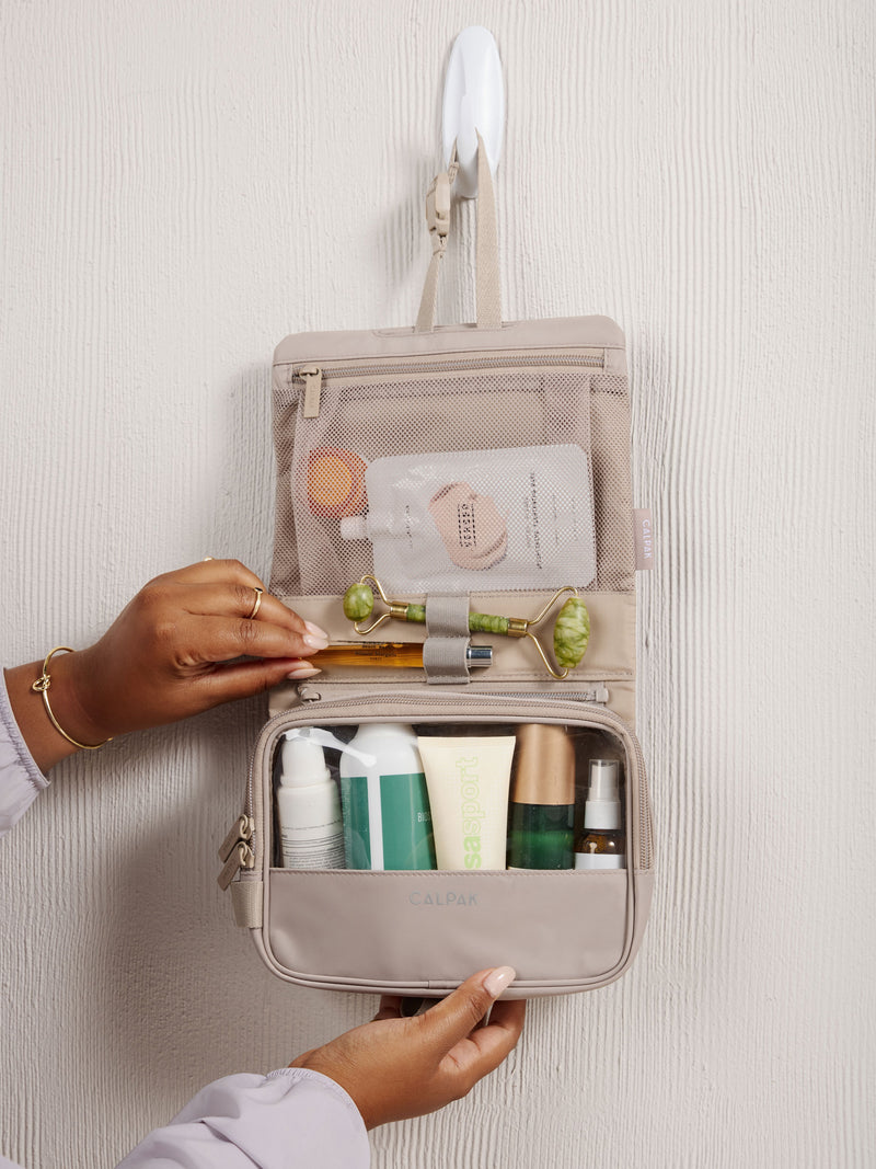 Model placing perfume bottle within elastic loops of CALPAK Fit gym toiletry bag full of toiletries in grayish beige