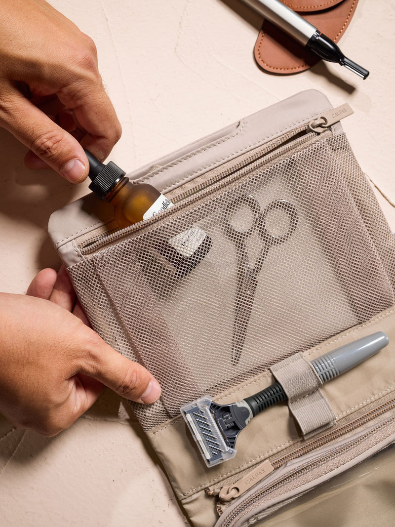 Model placing small toiletry item within interior mesh pocket of CALPAK Fit Hanging Dopp Kit in greige