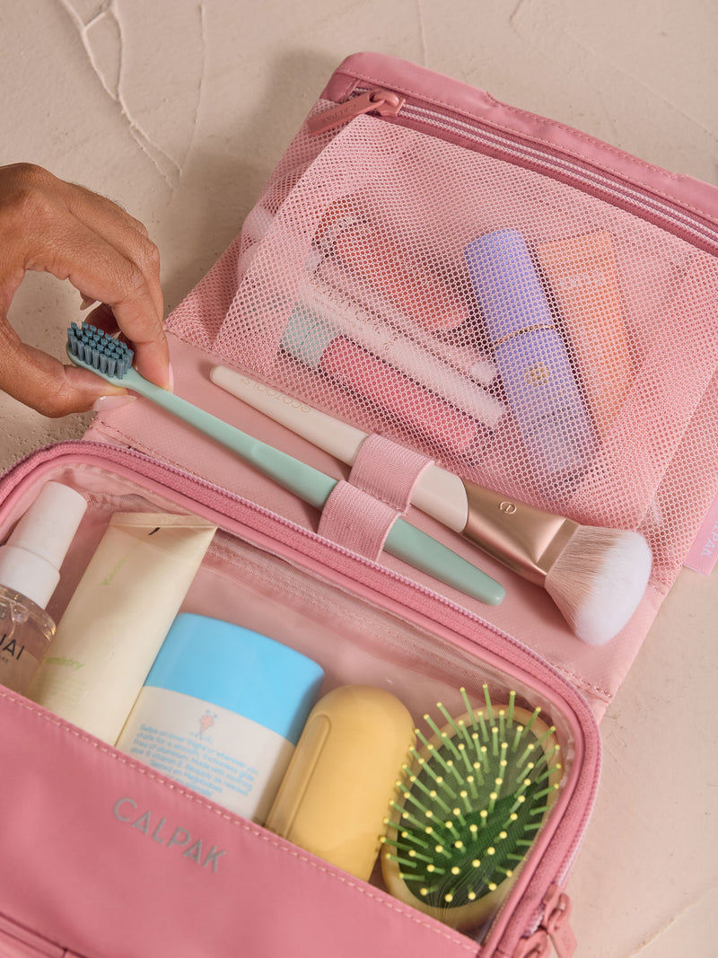 Model removing toothbrush from elastic loop of CALPAK Fit Hanging Dopp Kit in tea rose with toiletries packed within gym toiletry bag