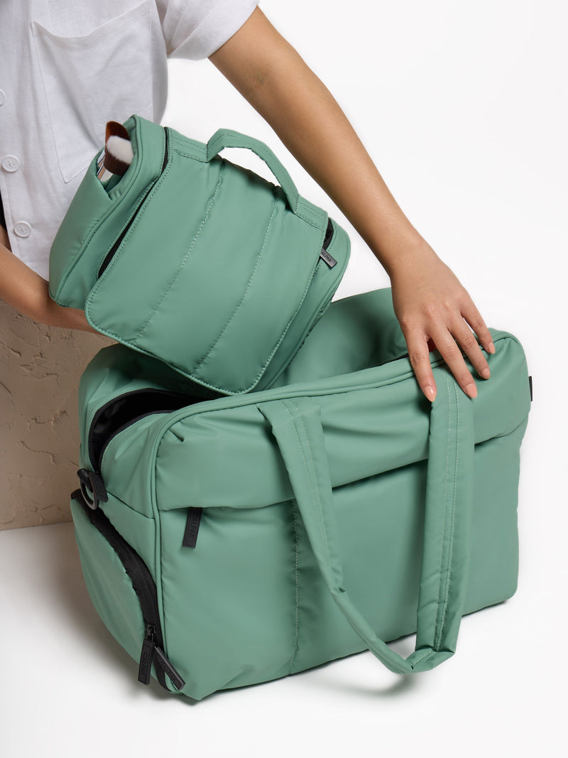 Model placing sage green CALPAK Hanging Toiletry Bag within Luka Duffel