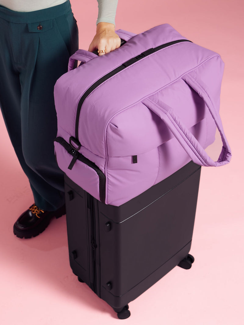 Model resting CALPAK Luka Large Duffel in lilac on top of CALPAK Hue Medium Carry-On Luggage in black