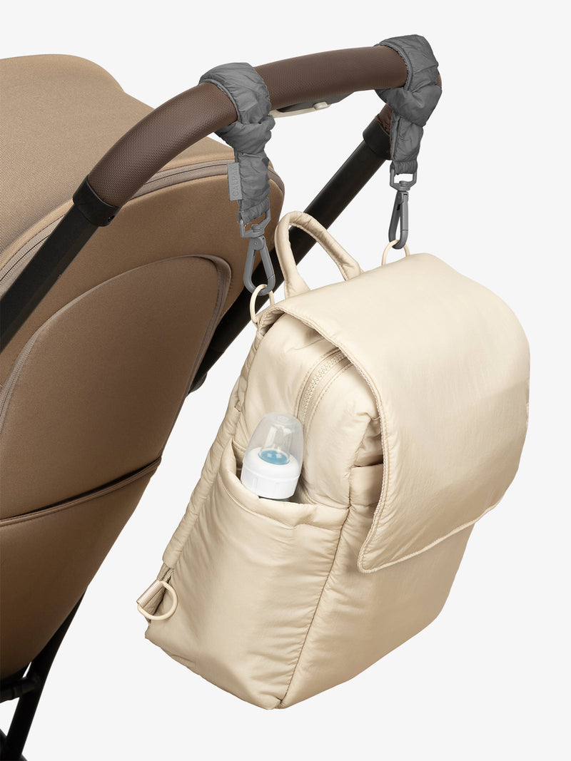 CALPAK Convertible Mini Diaper Backpack in white Oatmeal with grey Slate Stroller Straps attached to a brown stroller