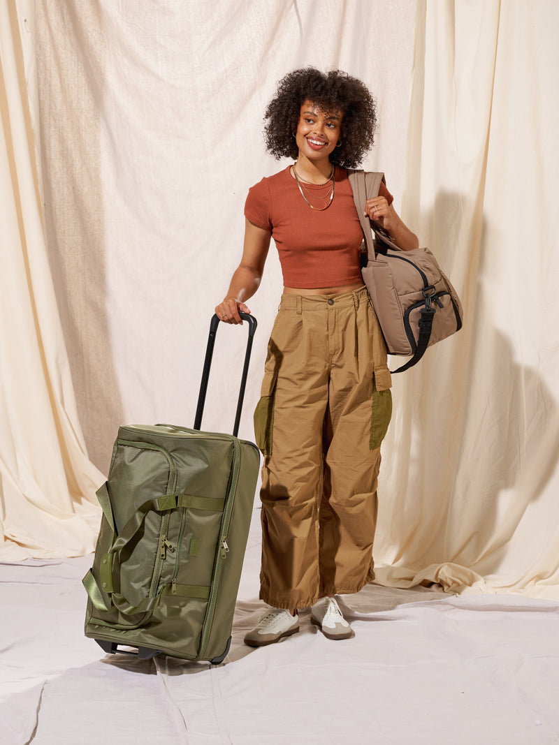 Model standing beside CALPAK Stevyn Large Rolling Duffle in moss while holding CALPAK Luka Duffel