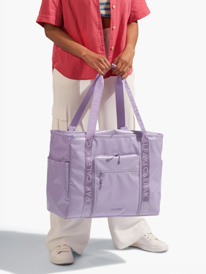 Model holding nylon tote straps of CALPAK Terra 35L Water Resistant Zippered Tote Bag in purple amethyst