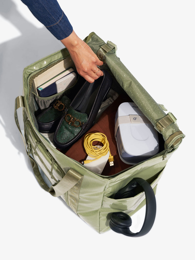 Model placing work shoes along with work essentials inside CALPAK Terra 35L Water Resistant Zippered Tote Bag in juniper