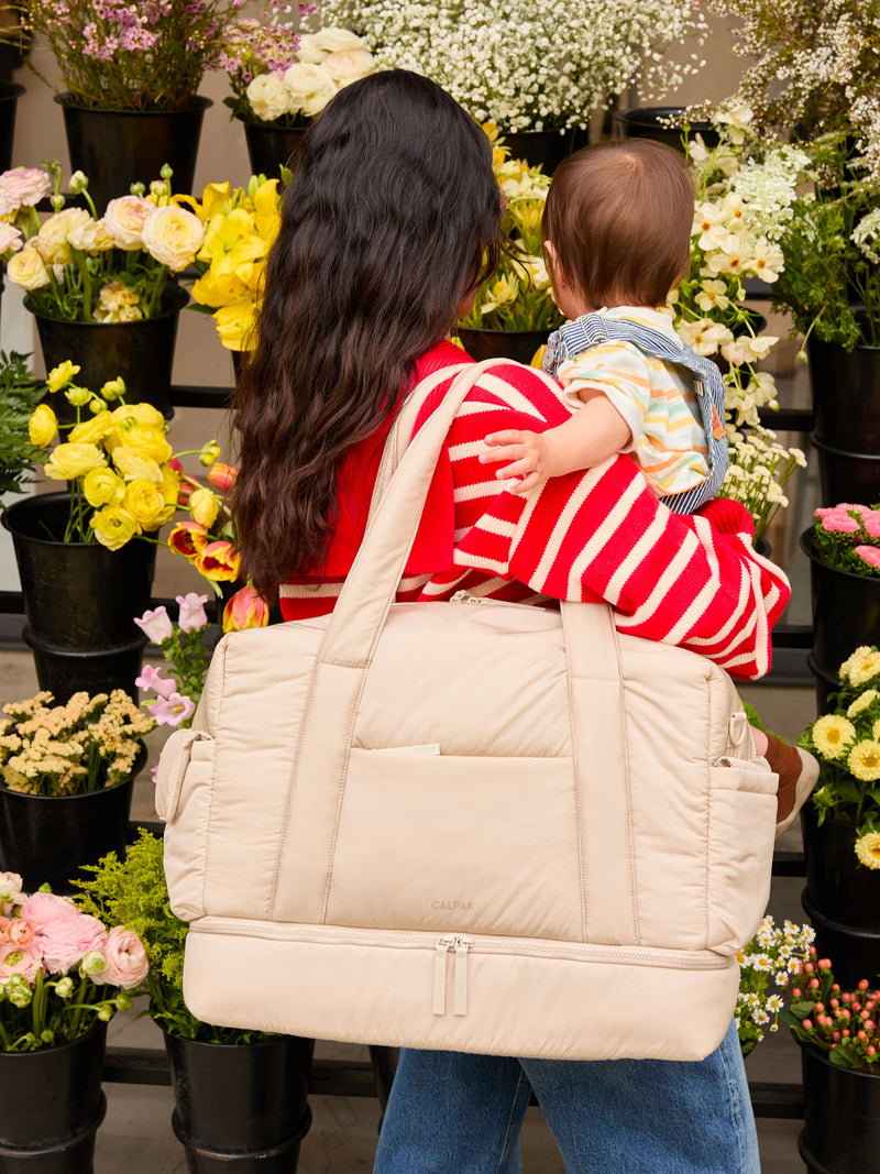 Model carrying baby and wearing the CALPAK Weekender Duffel in white Oatmeal